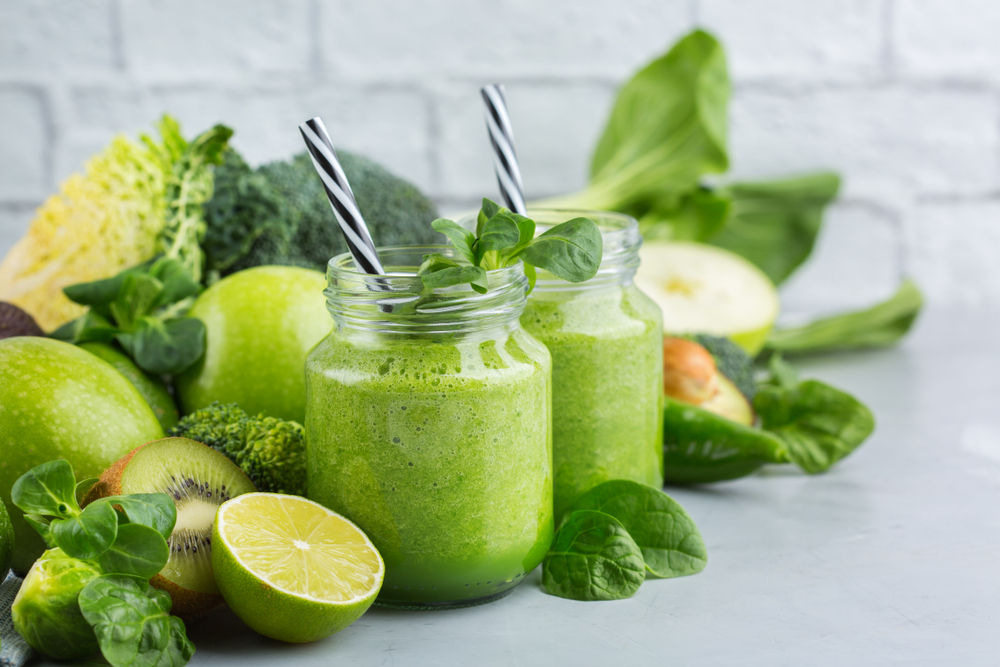 Green smoothies in jars sitting on a table with vegetables. 